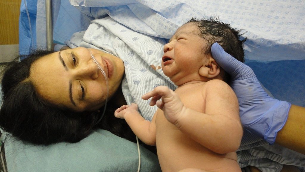 New born photo of Hispanic mom and baby during a c-section. Hand is holding up baby with full black hair to mom can see. Purpose illustrates that a c-section mom would benefit from a doula.