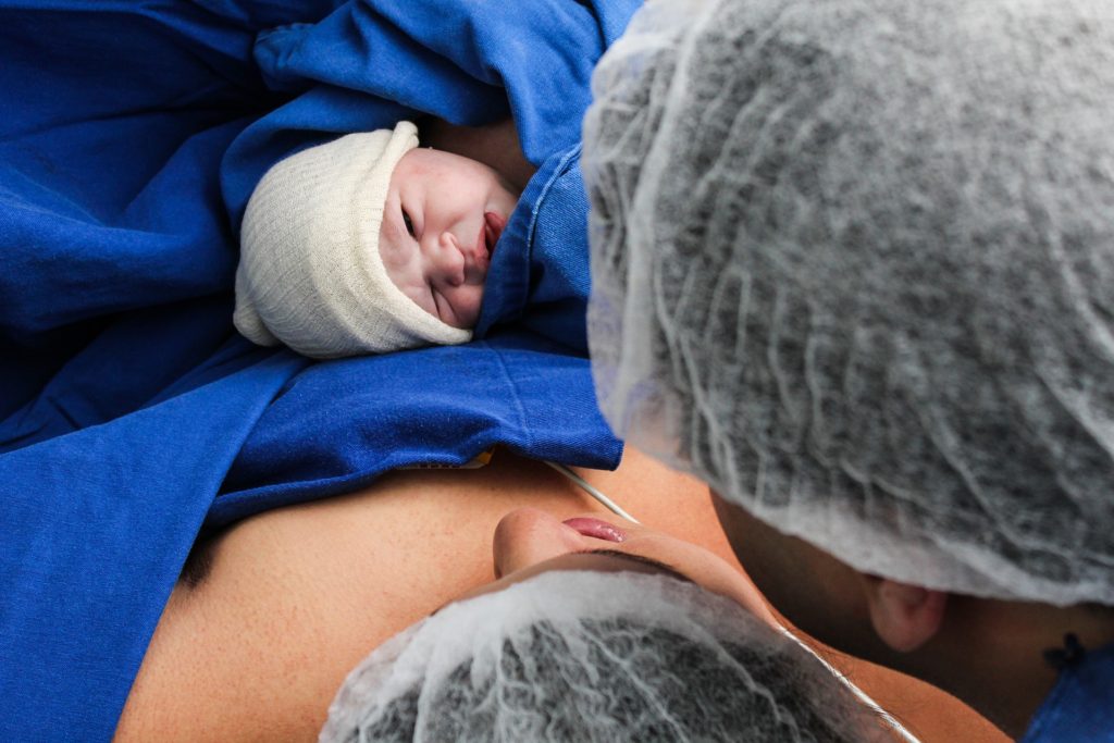 Photo of white woman with spouse looking down at new born baby. Baby is fair complicted and has a soft white baby cap on. The only face visible is of the baby. Mom and dad are wearing surgical caps. This photo is illustrating that even with a partner would benefit from a doula is a benefit.