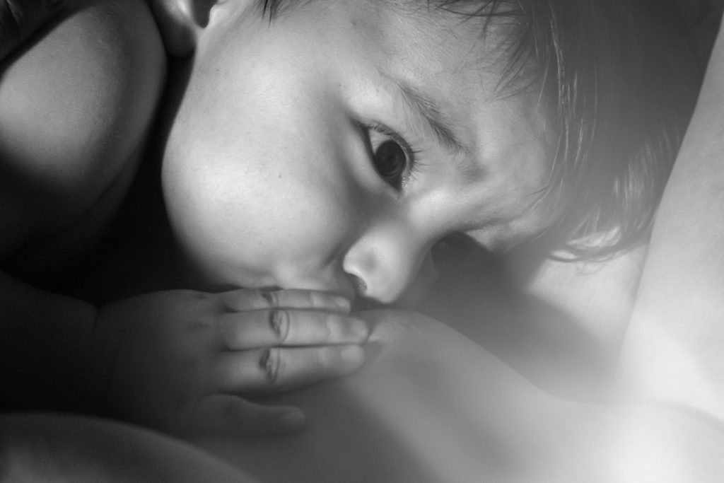 Black and white photo of baby breastfeeding. It is close up to baby and the breast. Baby's hand is resting close to his mouth while he nurses. Breastfeeding moms would benefit from a doula.