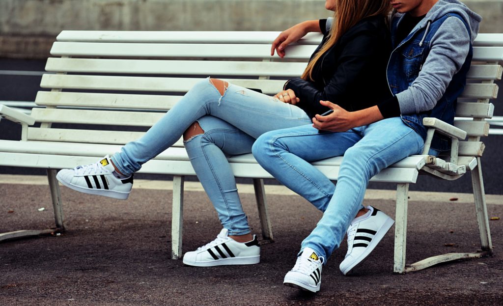 Couple snuggling on a park bench. Bother wearing jeans, a jacket, and matching tennis shoes. This is showing that a really easy, inexpensive date night idea is just enjoying each others company.