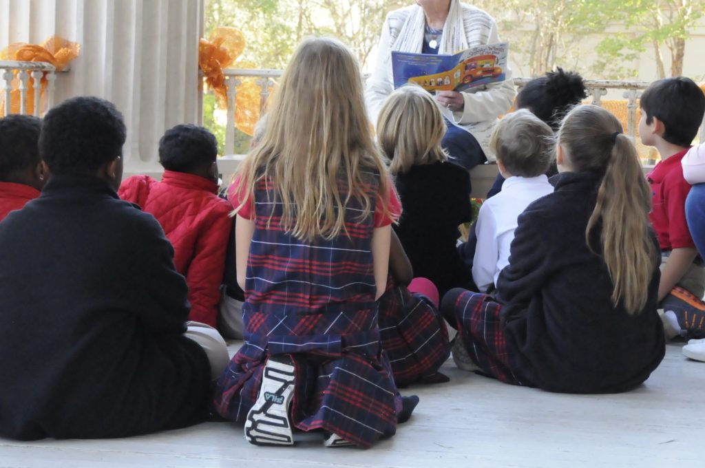 Elementray class sitting outside on a large paorch with an older women reading them a story. The picture is take to the backs of the students but the woman's face is not visible. Only the book. The book has a large yellow bus that covers the back and front of book. Orange mesh bows and tied onto the banister of the porch.