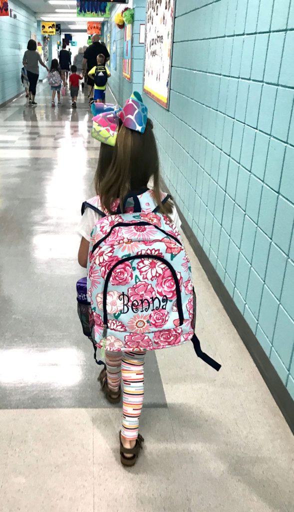 Little girl walking down a school hallway on her first day of kindergarten with a floral back pack with her name embroidered on the back and a big colorful bow in her fair. 