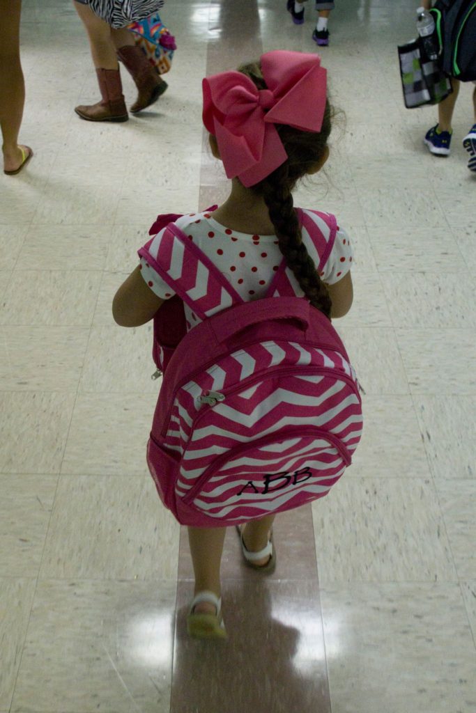 Little girl walking down the hallway on her first day of kindergarten with a pink and white stripe backpack with her monogram and a big pink bow in her hair.